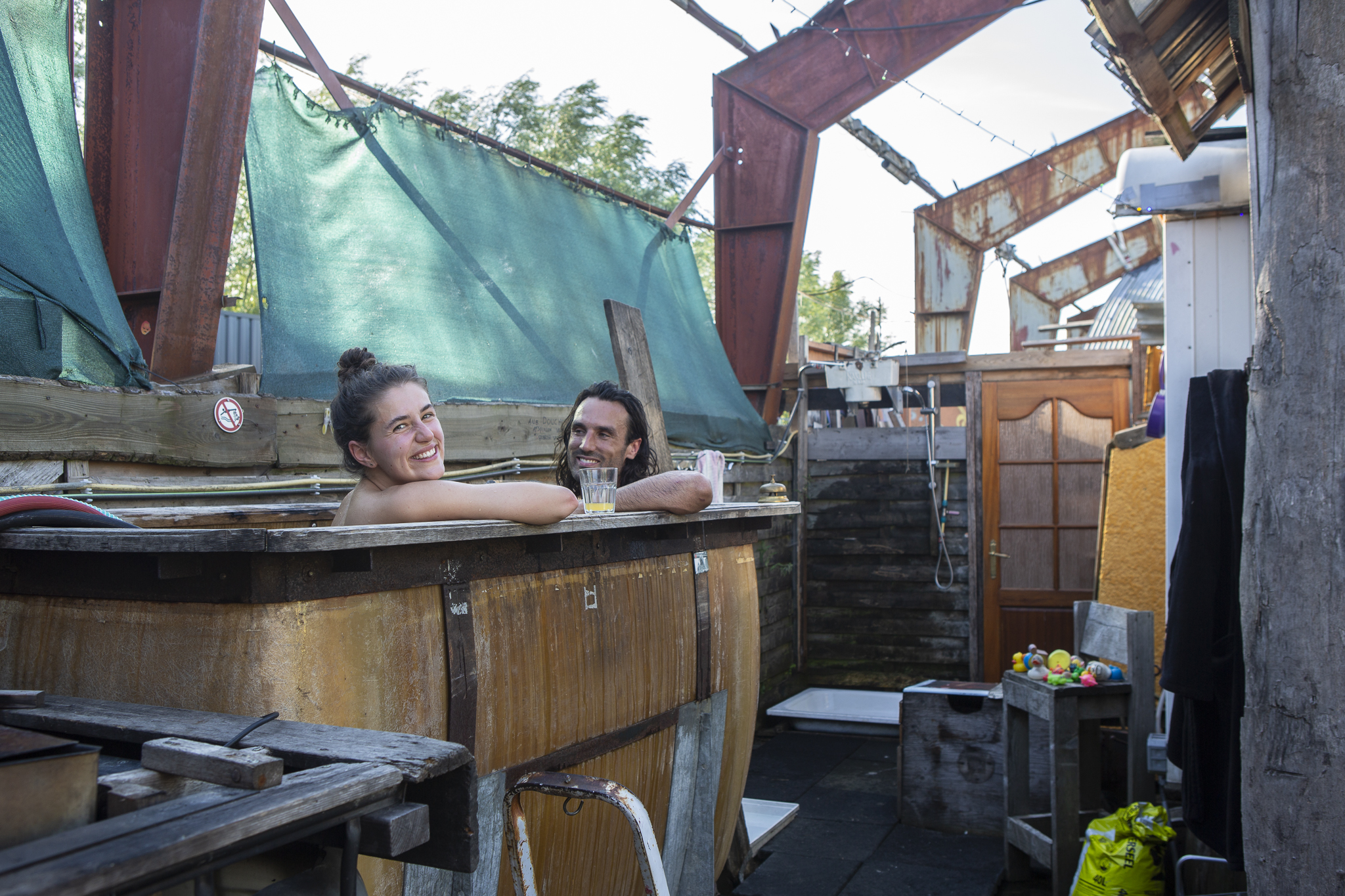 Twee mensen in de hottub, met een drankje. Industriële, creatieve achtergrond styling en een bak met badeendjes op de achtergrond.