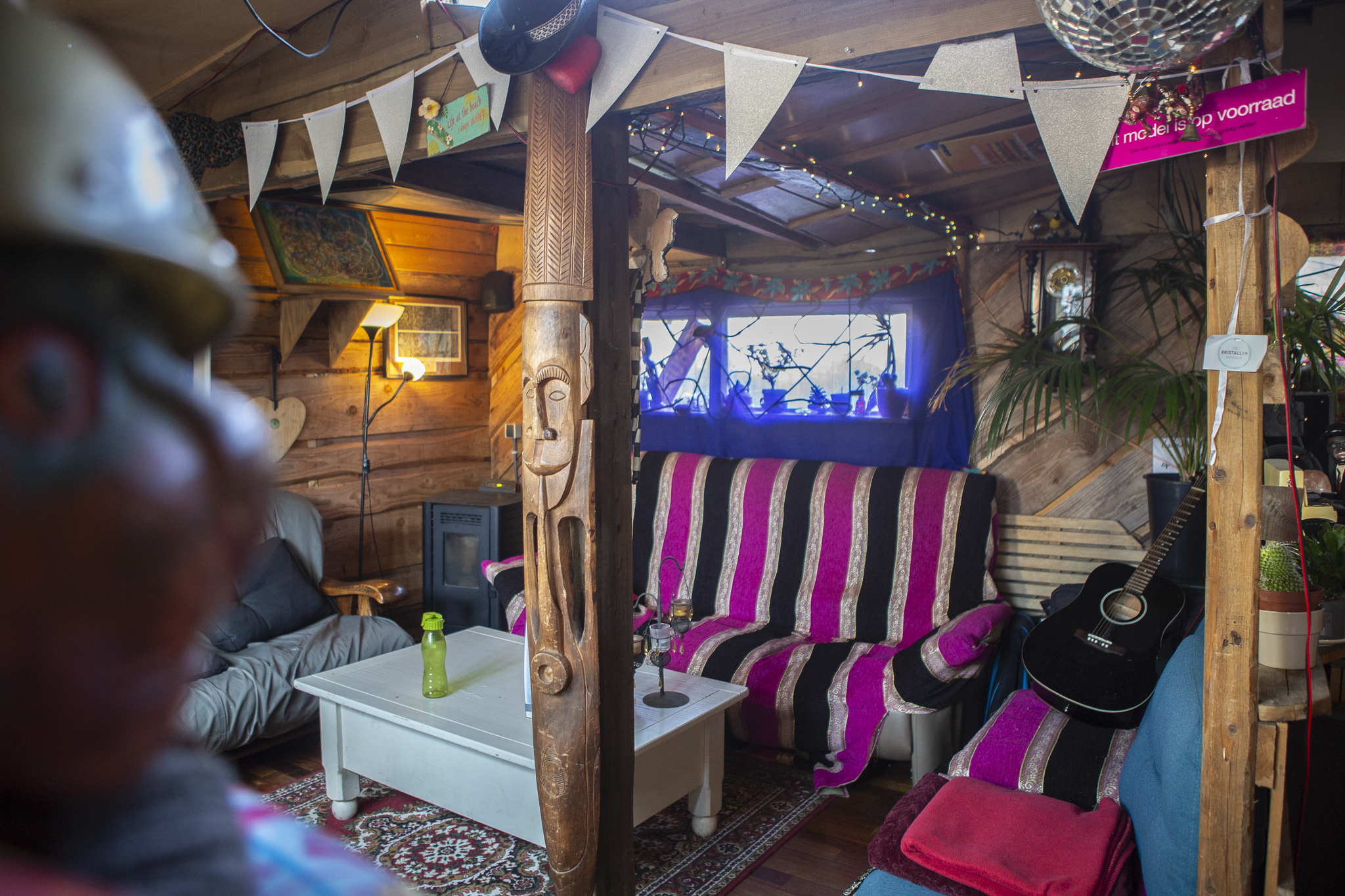 Relaxruimte met houten banken, kussens en kleedjes. Veel hout en felgekleurde, hippie stijl aankleding. Op een van de banken ligt een gitaar.