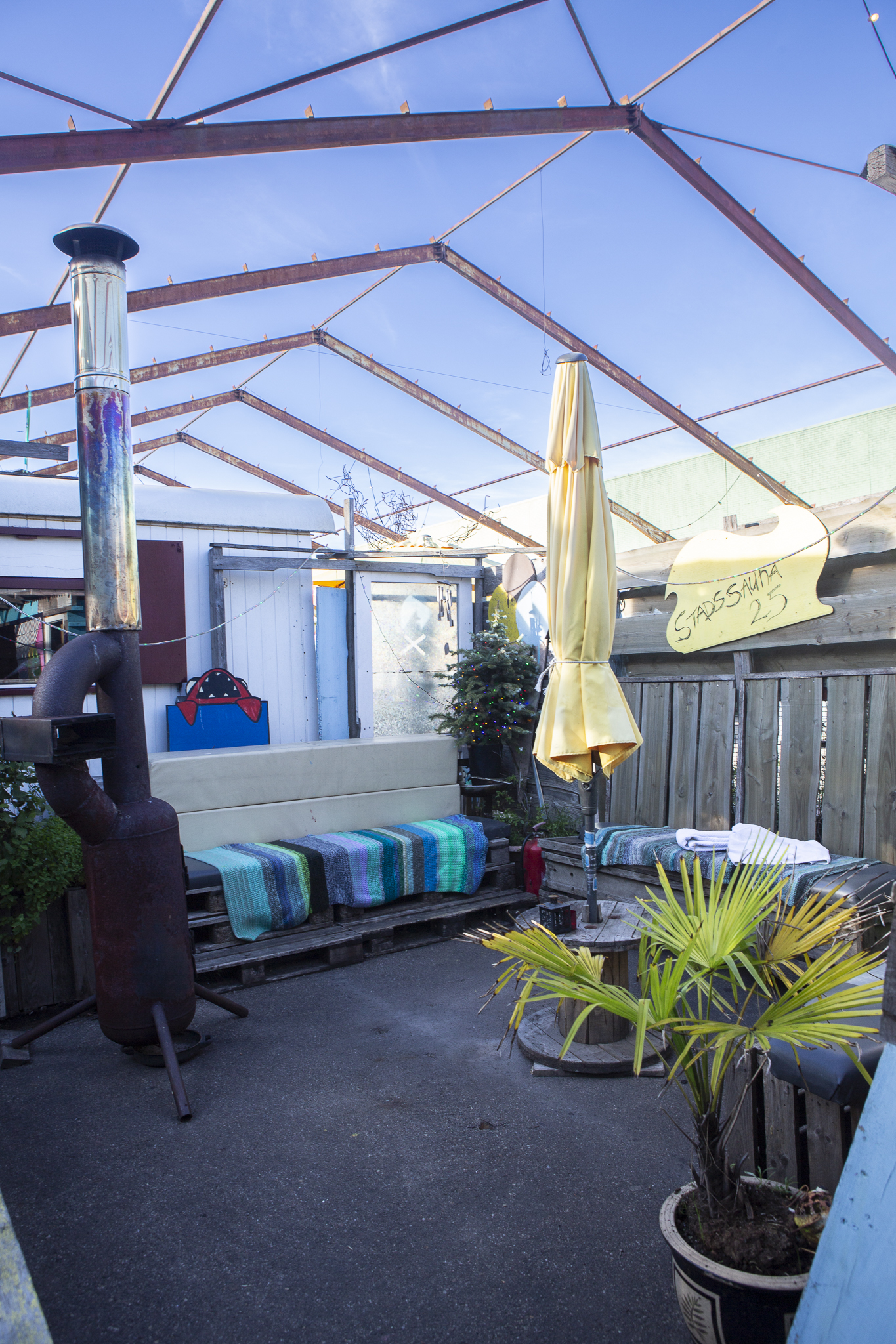 Lichte, blauwe relaxruime buiten. Industriele maar ontspannen look. Grote witte parasol en houtkachel aan weerzijden van de bank.